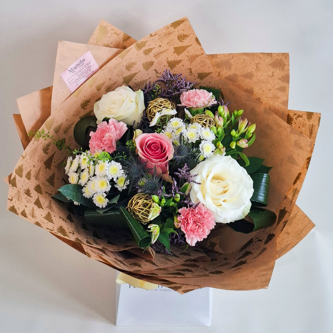 Frosted Blush Hand-Tied Bouquet - Magnolia the Florist The Frosted Blush Hand-Tied Bouquet offers a refined Christmas arrangement blending soft pink and white roses with delicate alstroemeria and white chrysanthemums. Eryngium and elegant gold accessories introduce subtle contrast and festive charm. Carefully wrapped in kraft paper and cellophane, this bouquet is presented in our signature boxes, making it a sophisticated and thoughtful gift for the holidays.  2
