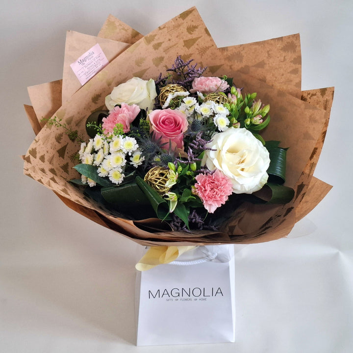 Frosted Blush Hand-Tied Bouquet - Magnolia the Florist The Frosted Blush Hand-Tied Bouquet offers a refined Christmas arrangement blending soft pink and white roses with delicate alstroemeria and white chrysanthemums. Eryngium and elegant gold accessories introduce subtle contrast and festive charm. Carefully wrapped in kraft paper and cellophane, this bouquet is presented in our signature boxes, making it a sophisticated and thoughtful gift for the holidays.  1