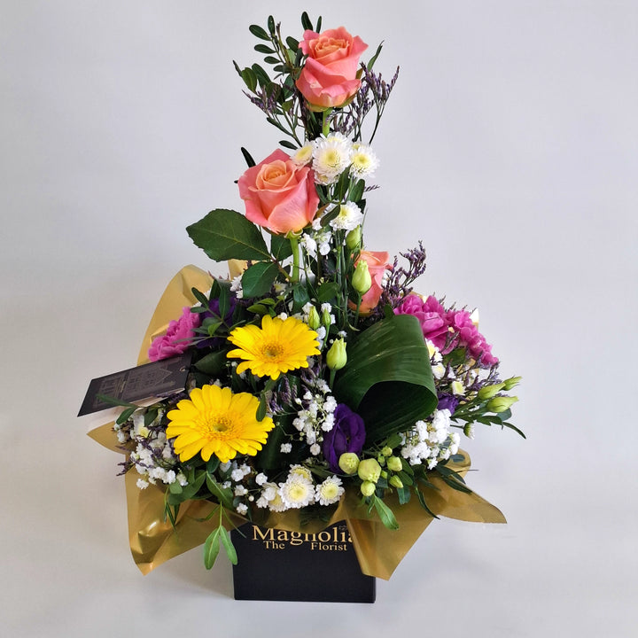 Barra Arrangement - Magnolia the Florist Barra is a warm and vibrant floral arrangement combining orange roses, germini, cerise carnations, white chrysanthemum, purple lisianthus, folded aspidistra, limonium, gyp and pistache. Presented in our Magnolia‑branded box and arranged in floral foam for long‑lasting freshness. For scale purposes, our branded box is 15cm (h) x 12cm (w) x 12cm (d), giving an overall size of approximately 35cm (h) x 18cm (w).  1