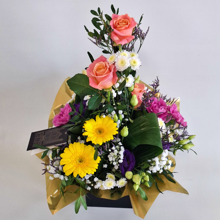 Barra Arrangement - Magnolia the Florist Barra is a warm and vibrant floral arrangement combining orange roses, germini, cerise carnations, white chrysanthemum, purple lisianthus, folded aspidistra, limonium, gyp and pistache. Presented in our Magnolia‑branded box and arranged in floral foam for long‑lasting freshness. For scale purposes, our branded box is 15cm (h) x 12cm (w) x 12cm (d), giving an overall size of approximately 35cm (h) x 18cm (w).  2