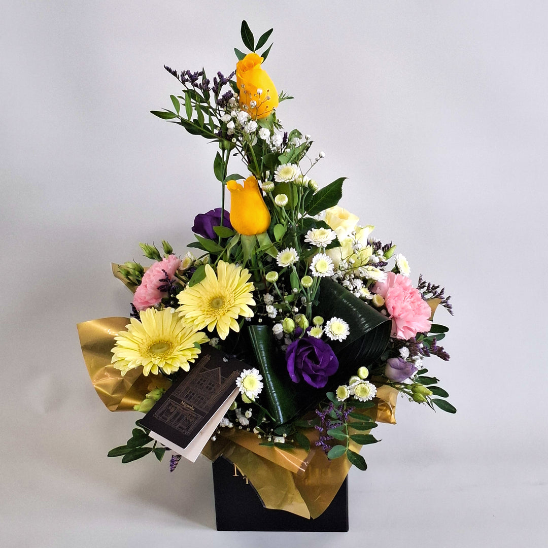 Cara Arrangement - Magnolia the Florist Cara is a joyful, sunshine‑inspired floral arrangement featuring yellow roses, yellow germini, pink carnations, white chrysanthemum, purple lisianthus, folded aspidistra, limonium, gyp and pistache. Presented in our Magnolia‑branded box and arranged in floral foam for long‑lasting freshness. For scale purposes, our branded box is 15cm (h) x 12cm (w) x 12cm (d), giving an overall size of approximately 35cm (h) x 18cm (w).  2