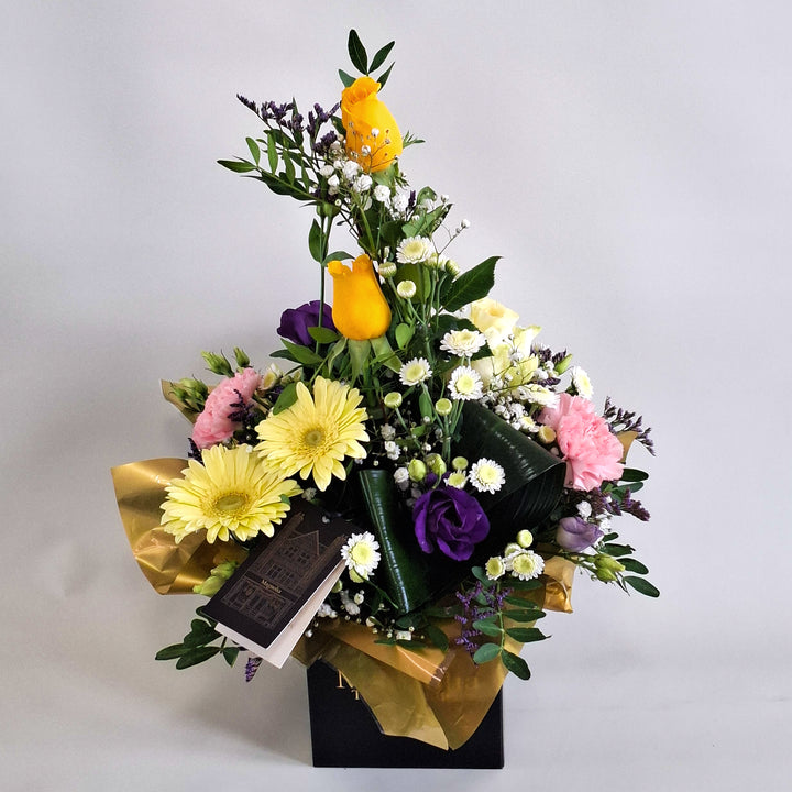 Cara Arrangement - Magnolia the Florist Cara is a joyful, sunshine‑inspired floral arrangement featuring yellow roses, yellow germini, pink carnations, white chrysanthemum, purple lisianthus, folded aspidistra, limonium, gyp and pistache. Presented in our Magnolia‑branded box and arranged in floral foam for long‑lasting freshness. For scale purposes, our branded box is 15cm (h) x 12cm (w) x 12cm (d), giving an overall size of approximately 35cm (h) x 18cm (w).  2
