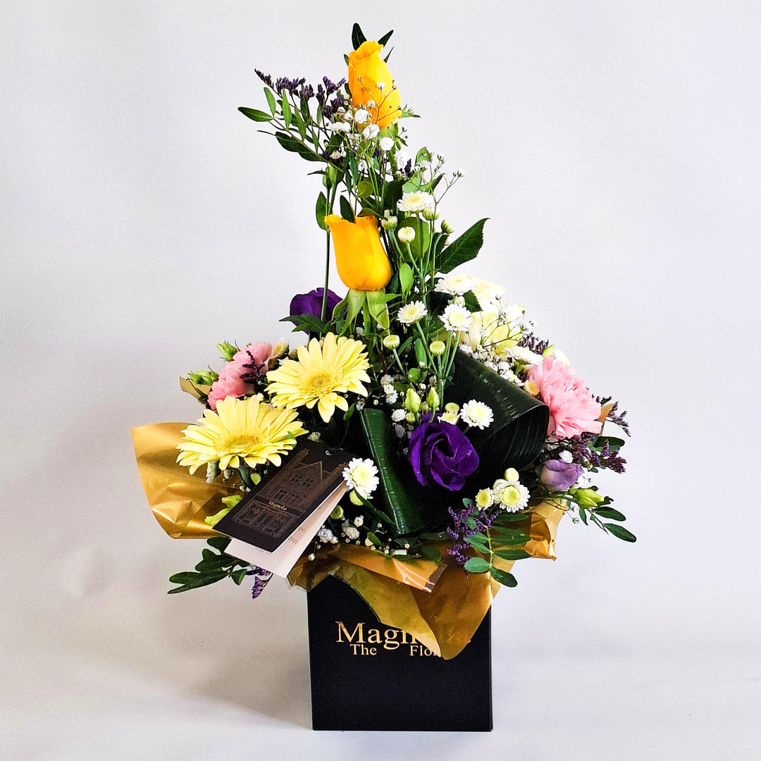 Cara Arrangement - Magnolia the Florist Cara is a joyful, sunshine‑inspired floral arrangement featuring yellow roses, yellow germini, pink carnations, white chrysanthemum, purple lisianthus, folded aspidistra, limonium, gyp and pistache. Presented in our Magnolia‑branded box and arranged in floral foam for long‑lasting freshness. For scale purposes, our branded box is 15cm (h) x 12cm (w) x 12cm (d), giving an overall size of approximately 35cm (h) x 18cm (w).  1
