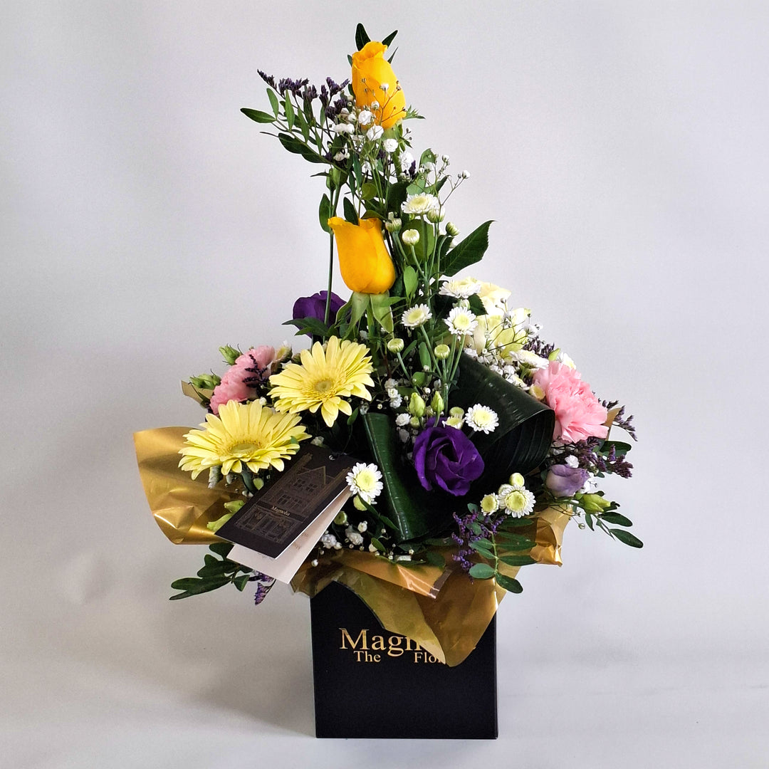 Cara Arrangement - Magnolia the Florist Cara is a joyful, sunshine‑inspired floral arrangement featuring yellow roses, yellow germini, pink carnations, white chrysanthemum, purple lisianthus, folded aspidistra, limonium, gyp and pistache. Presented in our Magnolia‑branded box and arranged in floral foam for long‑lasting freshness. For scale purposes, our branded box is 15cm (h) x 12cm (w) x 12cm (d), giving an overall size of approximately 35cm (h) x 18cm (w).  3