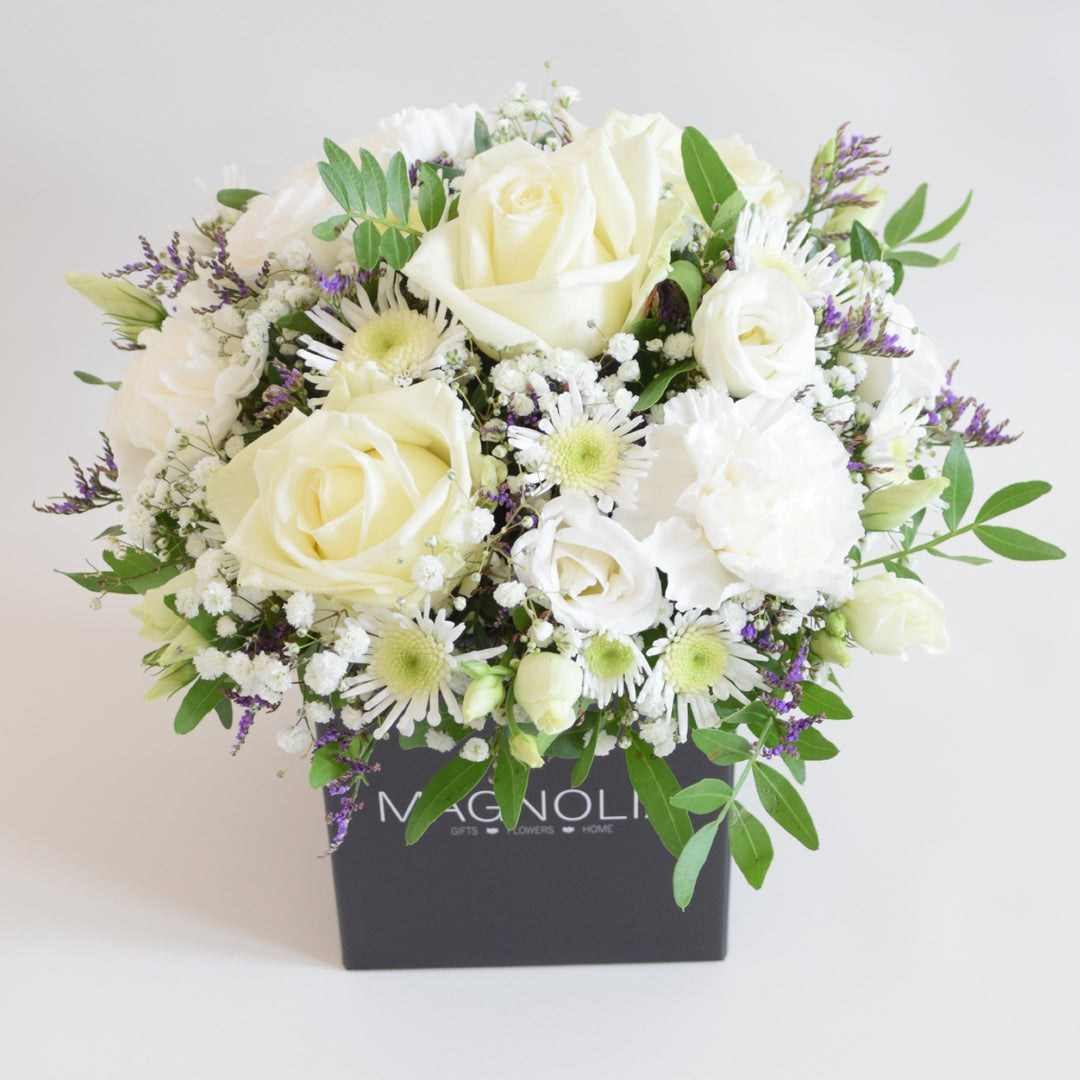 Dainty Kew Arrangement - Magnolia the Florist The Kew Arrangement from Magnolia the Florist, features a stunning mix of white florals with a hint of purple, including roses, carnations, lisianthus, gyp, chrysanthemum and limonium, all arranged in floral foam. Presented in a signature box, it makes a beautiful and sophisticated gift for any occasion. For scale purposes, the box container measures approximately 15cm x 12cm x 12cm, giving the overall arrangement size of 25cm h x 18cm w approximately.  1