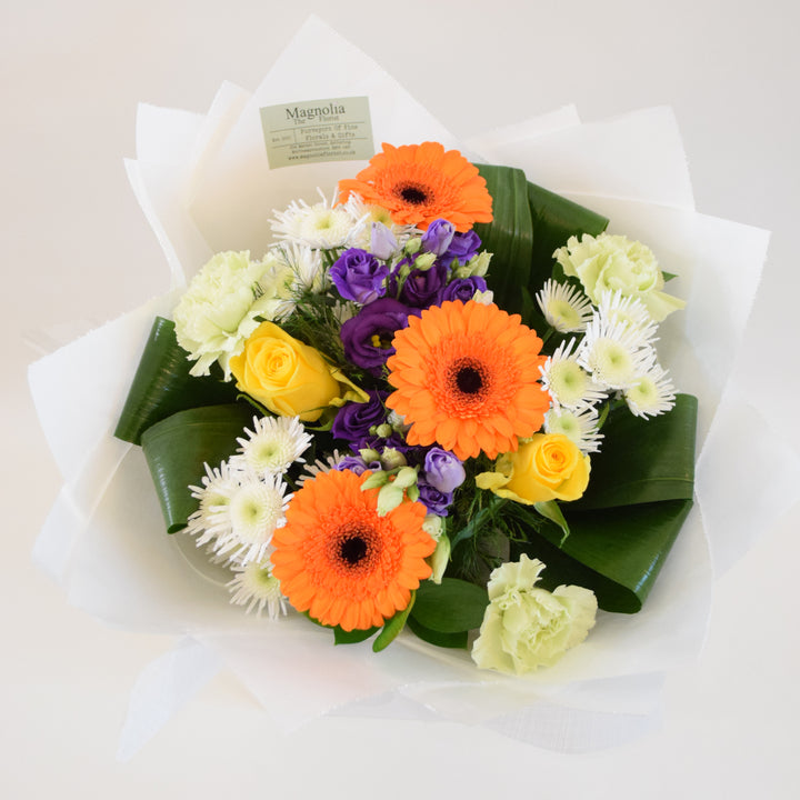 Savoy Hand-Tied Bouquet - Magnolia the Florist With vibrant hues of yellow, orange, green, and purple, the Savoy Hand-Tied Bouquet from Magnolia the Florist, is a stunning mix of Roses, Mini Gerbera, Lisianthus, Carnations, Chrysanthemums, and foliage. Expertly arranged in our signature box bags, this aqua bouquet is sure to brighten any space. Perfect for any occasion, bring a touch of nature&