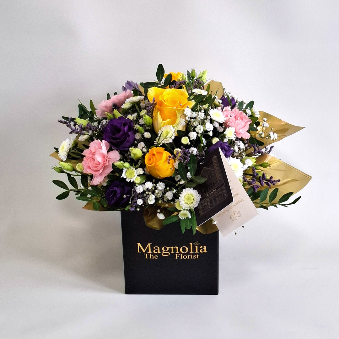 Dainty Somerset Arrangement - Magnolia the Florist A bright and uplifting floral arrangement designed to bring warmth into any space. Somerset features a joyful blend of yellow roses, pink carnations, white chrysanthemum, purple lisianthus, limonium, gyp and pistache. Presented in our Magnolia‑branded box and arranged in floral foam for long‑lasting freshness. For scale purposes, our branded box is 15cm(h) x 12cm(w) x 12cm(d), giving an overall size of approximately 25cm (h) x 18cm (w).  1