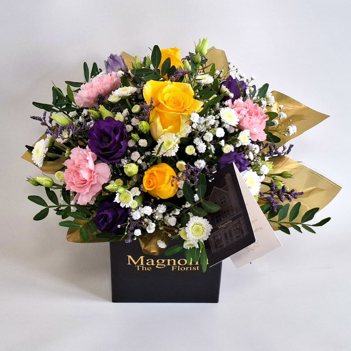 Dainty Somerset Arrangement - Magnolia the Florist A bright and uplifting floral arrangement designed to bring warmth into any space. Somerset features a joyful blend of yellow roses, pink carnations, white chrysanthemum, purple lisianthus, limonium, gyp and pistache. Presented in our Magnolia‑branded box and arranged in floral foam for long‑lasting freshness. For scale purposes, our branded box is 15cm(h) x 12cm(w) x 12cm(d), giving an overall size of approximately 25cm (h) x 18cm (w).  3