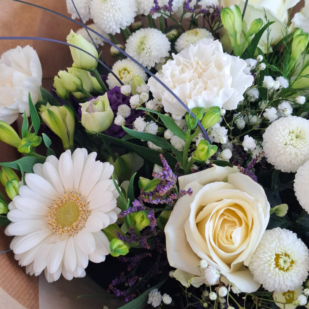 Wadenhoe Bouquet - Magnolia the Florist The Wadenhoe Bouquet is a meticulously hand-tied arrangement blending fresh Germini, Roses, Chrysanthemums, Eustoma, Alstroemeria, Carnations, Gypsophila, and Statice in elegant white and purple hues. Accented with lush greenery, it offers a sophisticated balance of texture and color, ideal for expressing refined taste and appreciation. Perfect for elevating any occasion with timeless floral beauty.  3