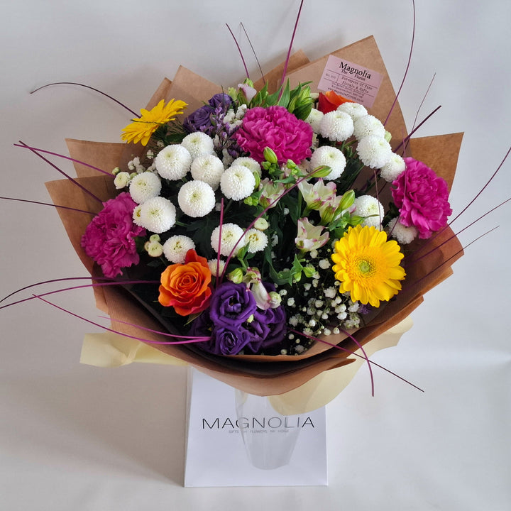 Yarwell Bouquet - Magnolia the Florist The Yarwell Bouquet is an expertly crafted hand-tied arrangement blending Roses, Germini, Chrysanthemums, Eustoma, Gyp, Alstroemeria, Carnations, and Statice. Its vibrant palette of yellow, orange, white, pink, cerise, and purple tones is beautifully complemented by fresh greenery, creating a harmonious and elegant floral display perfect for any occasion.  1
