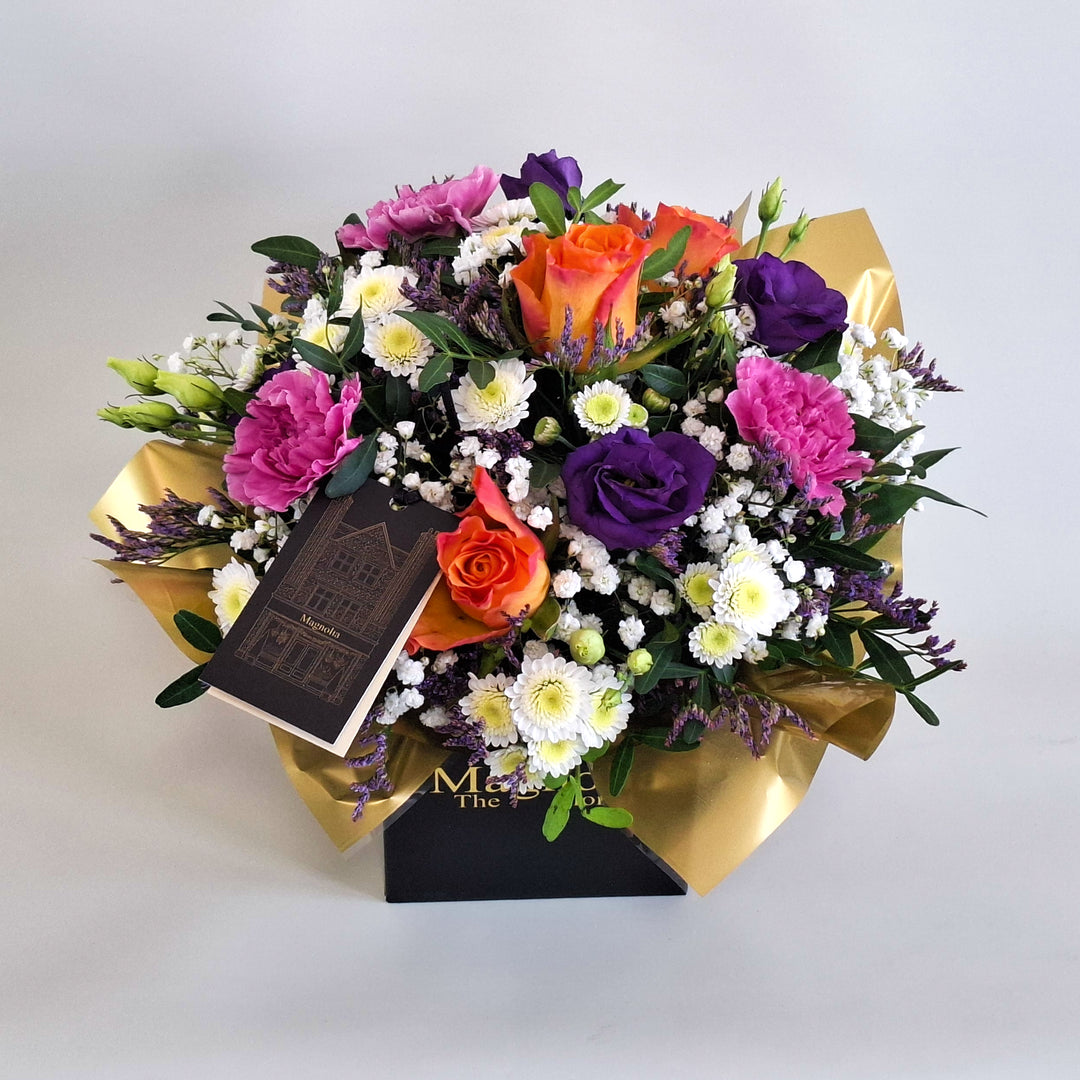 Petite York Arrangement - Magnolia the Florist York is a bold and vibrant floral arrangement featuring orange roses, cerise carnations, white chrysanthemum, purple lisianthus, limonium, gyp and pistache. Presented in our Magnolia‑branded box and arranged in floral foam for long‑lasting freshness. For scale purposes, our branded box is 15cm (h) x 12cm (w) x 12cm (d), giving an overall size of approximately 25cm (h) x 18cm (w).  3