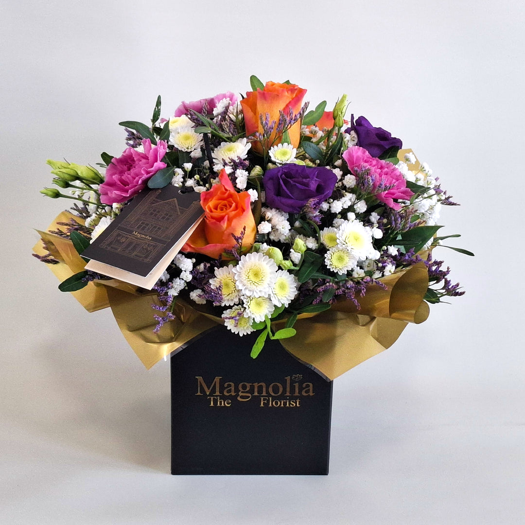Petite York Arrangement - Magnolia the Florist York is a bold and vibrant floral arrangement featuring orange roses, cerise carnations, white chrysanthemum, purple lisianthus, limonium, gyp and pistache. Presented in our Magnolia‑branded box and arranged in floral foam for long‑lasting freshness. For scale purposes, our branded box is 15cm (h) x 12cm (w) x 12cm (d), giving an overall size of approximately 25cm (h) x 18cm (w).  1