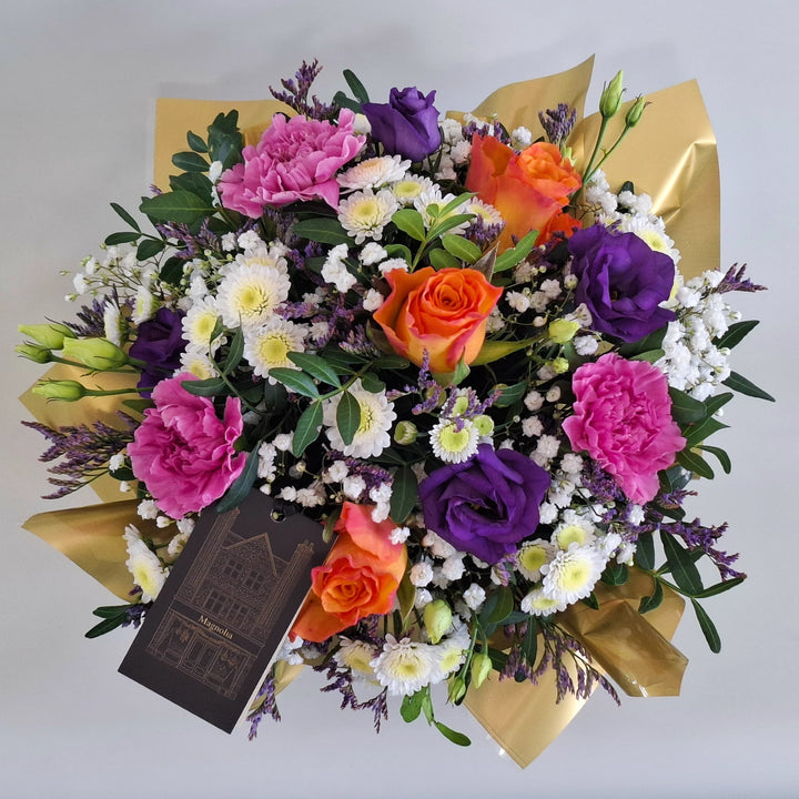 Petite York Arrangement - Magnolia the Florist York is a bold and vibrant floral arrangement featuring orange roses, cerise carnations, white chrysanthemum, purple lisianthus, limonium, gyp and pistache. Presented in our Magnolia‑branded box and arranged in floral foam for long‑lasting freshness. For scale purposes, our branded box is 15cm (h) x 12cm (w) x 12cm (d), giving an overall size of approximately 25cm (h) x 18cm (w).  2